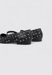 Zapatos planos negros de cuero con tachuelas plateadas, presentan una correa con hebilla. El diseño tiene una punta afilada y una suela plana.
