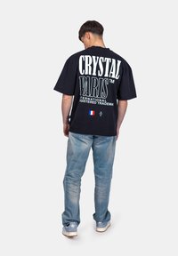 T-shirt noir surdimensionné avec un grand texte blanc "CRYSTAL PARIS", agrémenté d'une touche de drapeau français. À associer avec un jean bleu clair et des baskets.