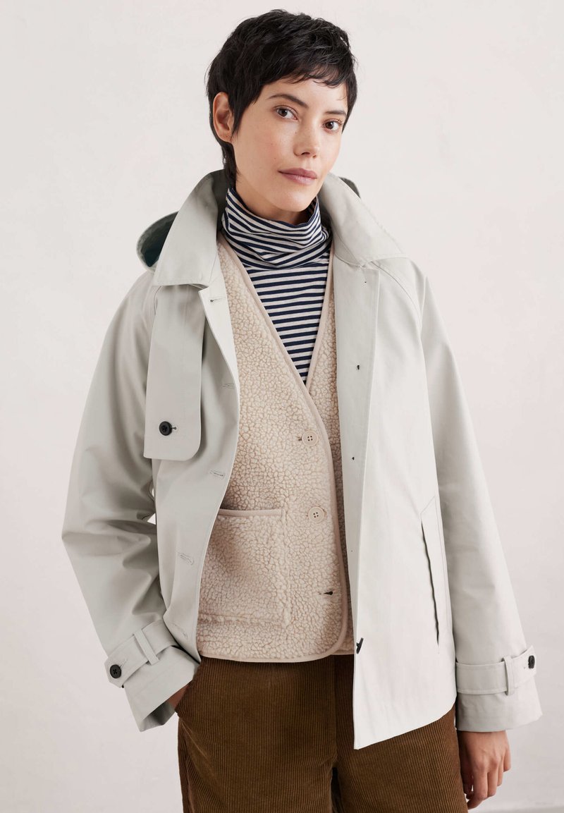 Vaaleanharmaa vedenpitävä takki, jossa on korkea kaulus, beigellä fleeceliivillä kerrostettuna, musta-valkoraidallinen poolopaita ja ruskeat corduroyhousut.