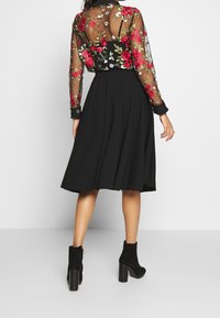 Donna che indossa una gonna midi nera, una blusa trasparente ricamata a fiori e stivaletti neri con tacco, in piedi contro uno sfondo neutro.