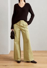 Schwarzer gerippter Pullover mit V-Ausschnitt, kombiniert mit weiten olivgrünen Hosen; Modell hält eine strukturiert braune Tasche, getragen mit spitzen schwarzen Schuhen.