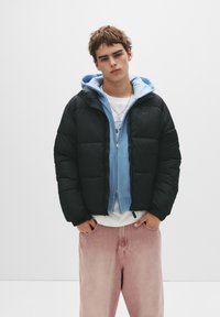 Sort pufferjakke med kviltet design, båret over en lyseblå zip-up hoodie og hvid skjorte, parret med lyserøde denim-bukser.
