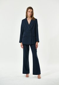 Blazer doppiopetto blu navy e pantaloni a palazzo abbinati in tessuto strutturato. Il blazer presenta grandi bottoni e due tasche frontali.