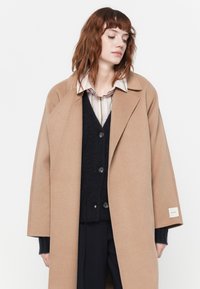 Cappotto di lana beige con ampio rever, vestibilità over size e tasche laterali, indossato sopra un maglione scuro e una camicia a righe chiare.