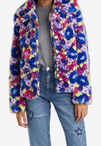 Kurtka ze sztucznego futra w żywym, wielokolorowym kwiatowym wzorze, z zamkiem błyskawicznym i szerokim kołnierzem. Noszona na szarej bluzce i podartych dżinsach.