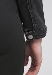 Veste en denim noire avec des coutures visibles, un bouton en métal et une silhouette slim, associée à un tissu noir uni en dessous.