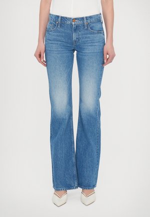 Kobieta ubrana w niebieskie dżinsy z wysokim stanem i rozkloszowanym dołem, z wyblakłymi udami, oraz w białe buty na obcasie o spiczastym nosku na jednolitym tle.