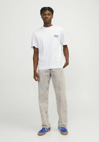 Camiseta blanca de algodón con mangas cortas, con texto azul. Combinada con jeans gris claro y zapatillas azules con acentos blancos.