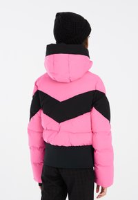 Veste matelassée rose et noire avec un motif en chevron, dotée d'une capuche et de larges manches. Un bonnet en tricot noir est porté sur la tête.
