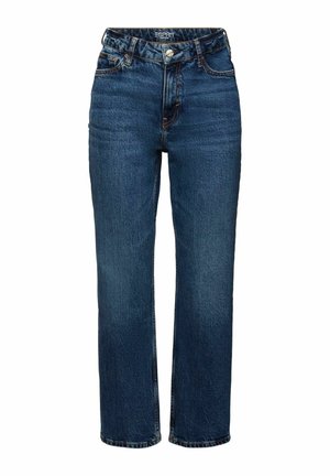 Personne portant un jean bleu, des bottes marron à lacets, une chemise verte boutonnée sous un pull rouge, tenant un petit sac à main marron.