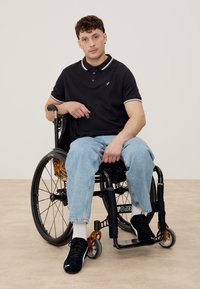 Άνδρας με κοντά σγουρά μαλλιά, ντυμένος με μαύρο πόλο και ανοιχτό μπλε τζιν, κάθεται σε αναπηρικό καροτσάκι σε ουδέτερο δάπεδο και φόντο.