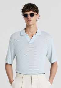 Mann in hellblauem Strick-Polo-Shirt, beigen, plissierten Hosen und quadratischen, schwarzen Sonnenbrillen, steht mit den Händen in den Taschen.