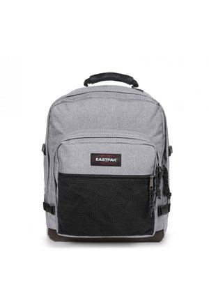 Mochila de tela gris con un bolsillo frontal de malla negra texturizada, asa superior y correas de hombro ajustables. Presenta un parche con el logo de Eastpak.