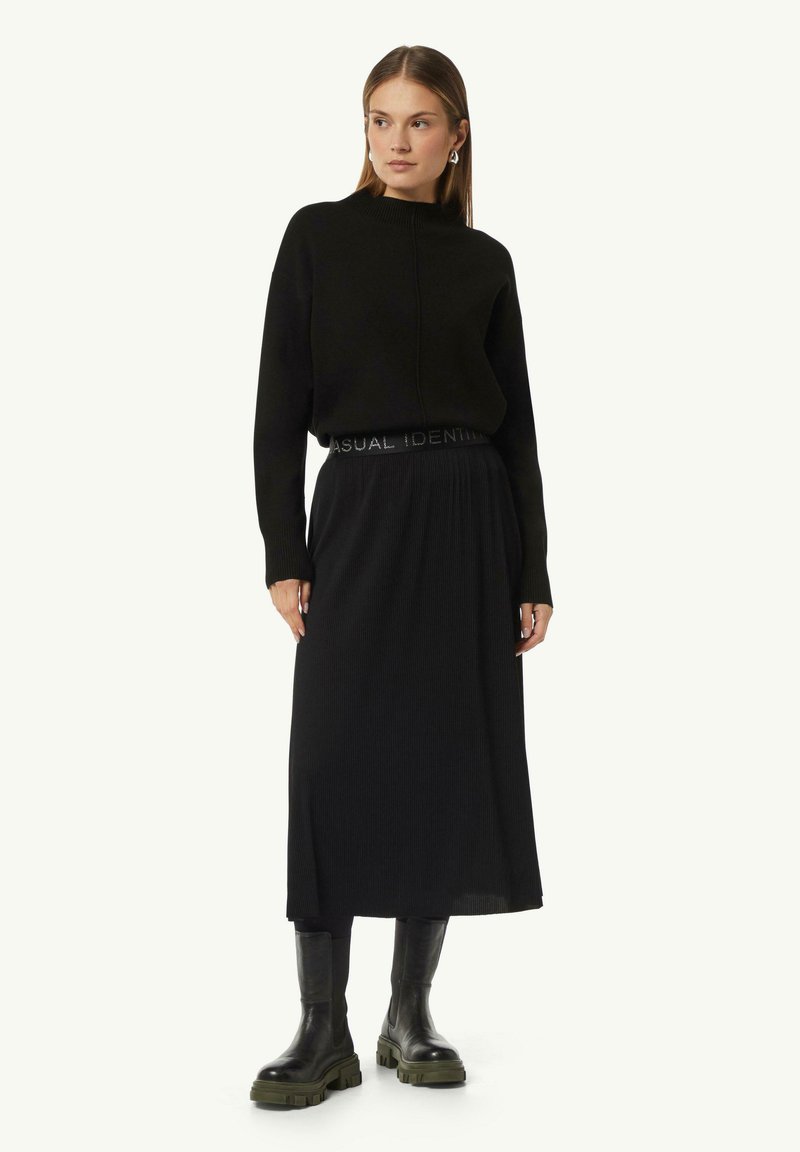 Black knit sweater with high neckline and ribbed texture. Midi-length black skirt with an elastic waistband featuring "CASUAL IDENTITI." Chunky black ankle boots.