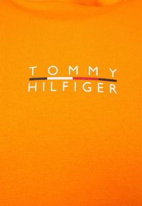 Pomarańczowy t-shirt z krótkim rękawem o prążkowanej fakturze, z logo "TOMMY HILFIGER" w białe, czerwone i niebieskie poziome paski.