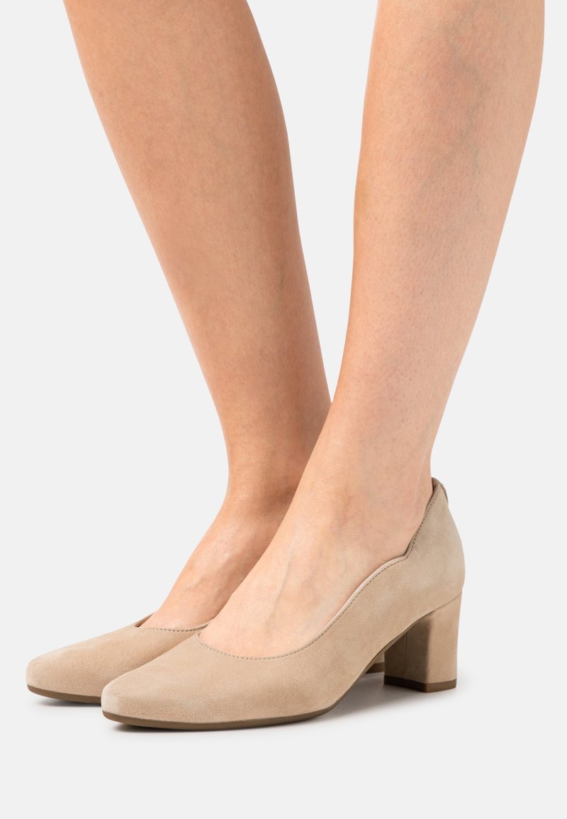 Pieds portant des chaussures en daim beige à petit talon bloc avec des bouts arrondis sur un fond clair.