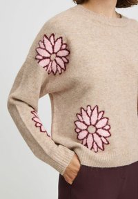 Pull en laine beige avec des poignets côtelés, orné de grandes broderies florales roses et bordeaux sur les manches. Texture douce et coupe décontractée.