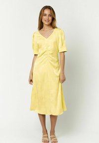 Gelbes Satin-Kleid mit V-Ausschnitt, kurzen Puffärmeln, gerafften Details an der Brust und einem fließenden, knielangen Saum. Glatte Textur.
