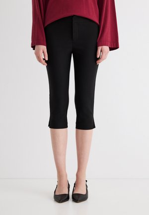 Άτομο που φοράει μαύρο στενό παντελόνι μέχρι τον αστράγαλο, μαύρα μυτερά παπούτσια και μακρυμάνικη μπορντό μπλούζα, στέκεται μπροστά σε λευκό απλό φόντο.