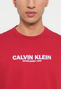 Κόκκινη βαμβακερή μπλούζα με λευκό κείμενο "CALVIN KLEIN" και "ΙΔΡΥΘΗΚΕ 1968" από κάτω σε έντονη, μοντέρνα γραμματοσειρά. Στρογγυλή λαιμόκοψη.