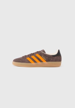 Hnedý semišový Adidas Gazelle teniska s oranžovými pruhmi, gumovou podrážkou a čiernou pätovou záložkou, zobrazená z bočného profilu na bielom pozadí.
