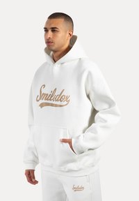 Sudadera blanca con un gran logo beige de "Smilodox", bolsillo tipo canguro, puños y bajo acanalados, combinada con pantalones blancos a juego.