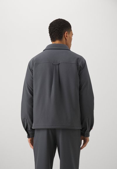 Gray lightweight jacket with a collared neck, long sleeves, and a buttoned cuff detail. Back features a central pleat for added movement.
