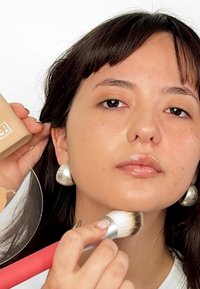 Malli pitää nestemäistä meikkipohjaa kädessään; hän levittää meikkipohjaa siveltimellä. Hopeiset rengaskorvakorut, luonnollinen ihon rakenne ja kiiltävät huulet näkyvät.