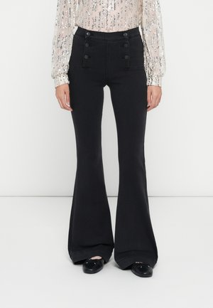 Vrouw draagt zwarte wijde broek met hoge taille, zes knopen aan de voorkant en zwarte ballerina's, gecombineerd met een wit lang mouw pailletten top.