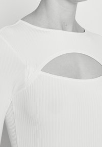 Vit ribbad långärmad topp med rundad halsringning och ett utskuret detalj vid bröstet, tillverkad av ett stretchigt material.