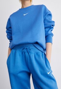 Person som bär en blå Nike-träningsoverall med vikta ärmar, händerna i fickorna, och vit Nike swoosh-logga på tröjan och byxorna.