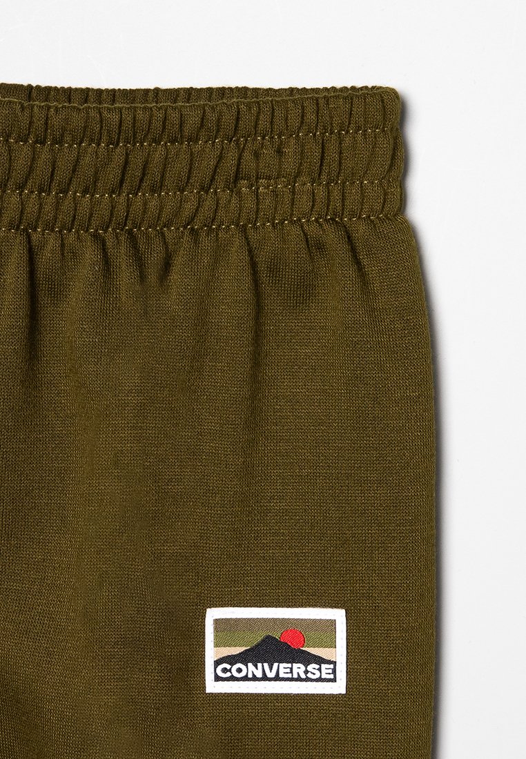 Jogging olive en vert avec une taille froncée et un petit patch rectangulaire blanc arborant un design de montagne noire et de soleil rouge.
