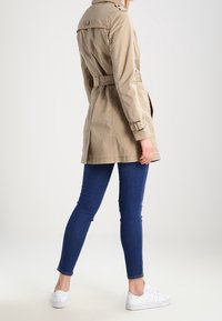 Beige trenchcoat met een ceintuur in de taille, knoopdetails op de schouders en lange mouwen, gedragen over blauwe jeans en witte sneakers.