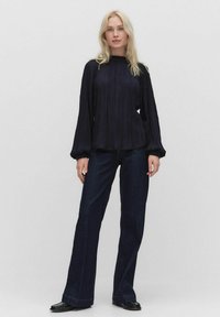 Marineblå plisseret bluse med høj halsudskæring og lange ballonærmer, parret med mørke denim flared bukser og sorte sko.