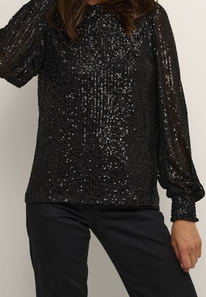 Femme portant un haut noir à manches longues pailleté avec manches transparentes et un pantalon noir, posant avec une main sur la hanche.