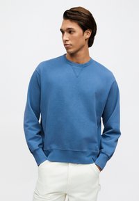 Νεαρός άντρας με καστανά μαλλιά, φορώντας μπλε φούτερ με στρογγυλή λαιμόκοψη και λευκό παντελόνι, με χέρια στις τσέπες, κοιτάζει προς τα πλάγια σε απλό φόντο.