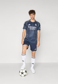 Tamsiai mėlyna futbolo marškinėliai su baltais akcentais, turintys Real Madrid logotipą. Su derančiais tamsiai mėlynais šortais, baltais kojinėmis ir sportbačiais, stovintys ant futbolo kamuolio.