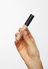 Vloeibare concealer in een doorzichtige tube met een zwarte dop, vastgehouden door een hand. Het product is lichtbeige met gedrukte branding aan de zijkant.