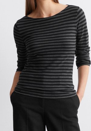 Kobieta ubrana w dopasowaną czarno-ciemnoszarą koszulkę w poziome paski z długim rękawem i czarne spodnie, ręce w kieszeniach, na jednolitym tle.