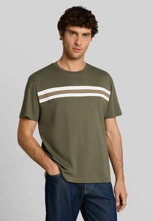 Homme aux cheveux bouclés foncés porte un t-shirt vert à manches courtes avec des rayures blanches et marron sur la poitrine et un jean bleu, debout avec la main dans la poche.