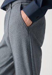 Pilki houndstooth rašto kelnių dizainas, pritaikytas, su šoninėmis kišenėmis ir lygia priekiu, derinami su tamsiai mėlyna marškiniais.