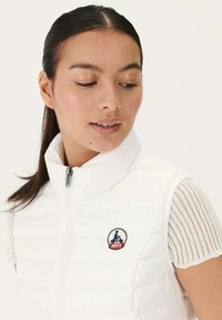 Chaleco de plumas blanco con cuello alto y cremallera frontal, que presenta un parche redondo con el logo en el lado izquierdo del pecho y un patrón acolchado.