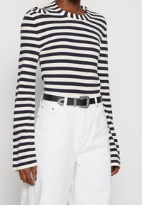 Navy und weiß gestreiftes Langarmoberteil mit einem Bootsausschnitt, kombiniert mit hochgeschnittenen weißen Hosen und einem schwarzen Gürtel mit einer silbernen Schnalle.