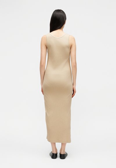 Γυναίκα με μακριά μαύρα μαλλιά στέκεται γυρισμένη προς τα πίσω, φοράει μπεζ μακρύ αμάνικο φόρεμα και μαύρα επίπεδα παπούτσια σε λευκό απλό φόντο.