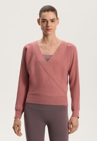 Pink wrap knit sweater with ribbed texture, deep V-neck, long sleeves, and fitted hem. Worn over a matching tank top and leggings.