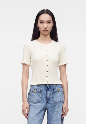 Femme aux longs cheveux noirs portant un haut crème côtelé à manches courtes avec boutons et un jean bleu taille haute avec de grandes poches avant.