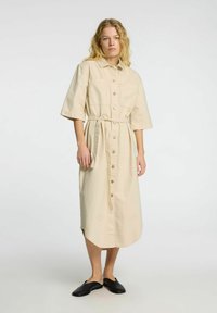 Beige skjortekjole med korte ærmer, bælte i taljen, siden slids og to forlommer. Sammen med sorte slip-on sko.