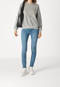 Sudadera gris con detalles de encaje, puños y dobladillo acanalados, combinada con jeans ajustados azules y zapatillas blancas. Bolsa negra colgada del hombro.