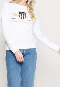 Bílá mikina s výšivkou loga, která obsahuje červený a modrý štít, zlaté doplňky a klasický kulatý výstřih; doplněná modrými džínami.