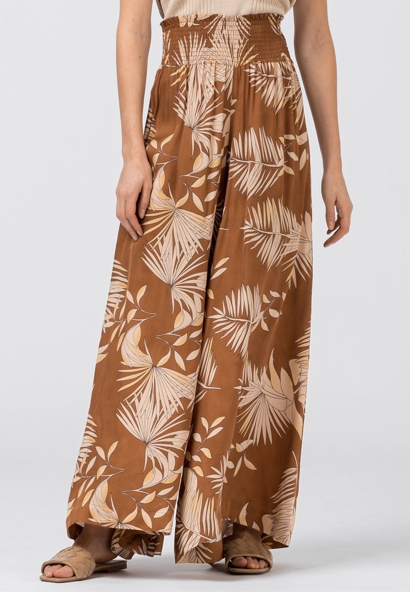 Hnědé široké kalhoty s nařaseným pasem, zdobené světle béžovým tropickým vzorem listů. Vyrobené z měkké tkaniny, s volným střihem.